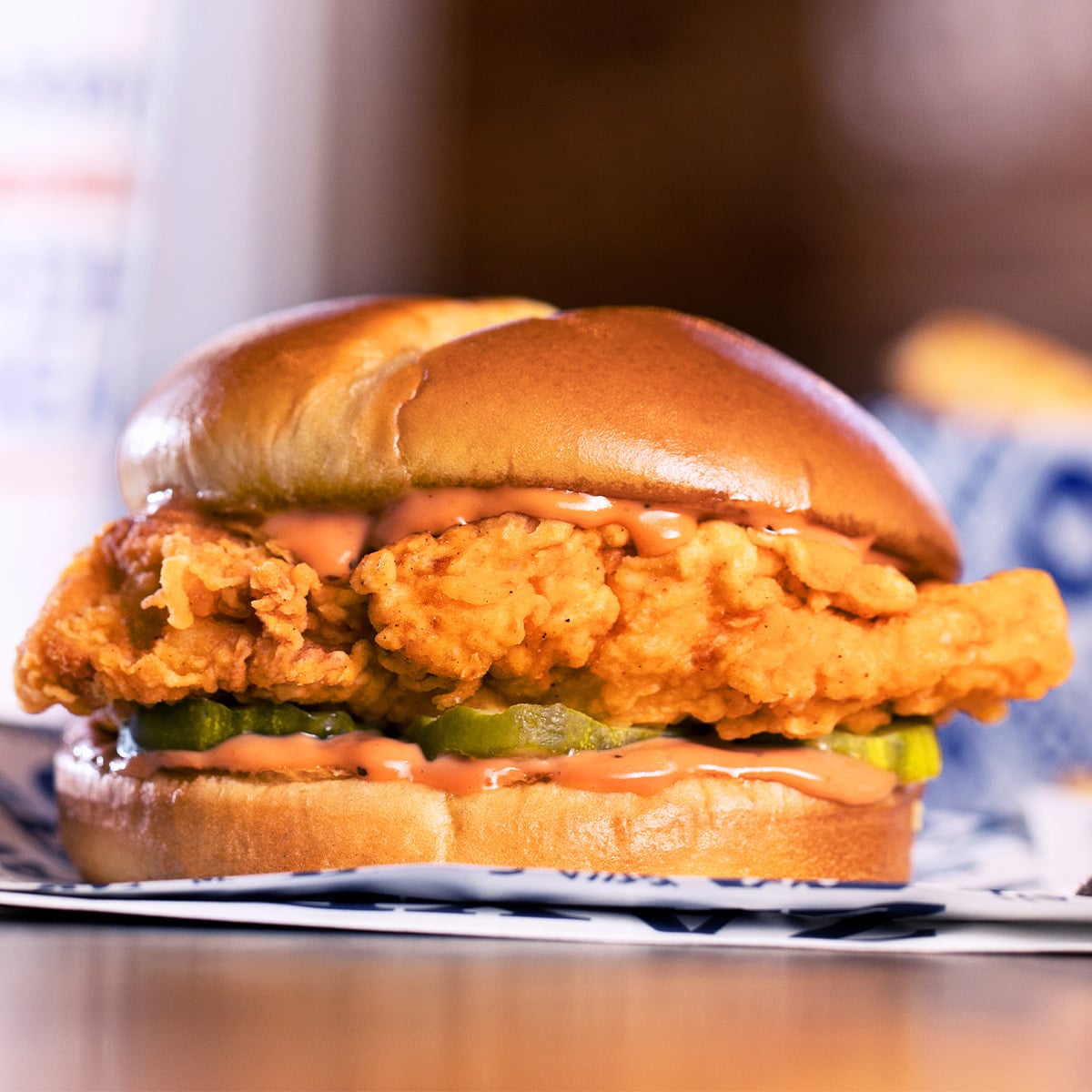 A close-up of a crispy Chicken Sandwich with pickles and a creamy orange sauce on a shiny, toasted bun, sitting on a paper wrapper.