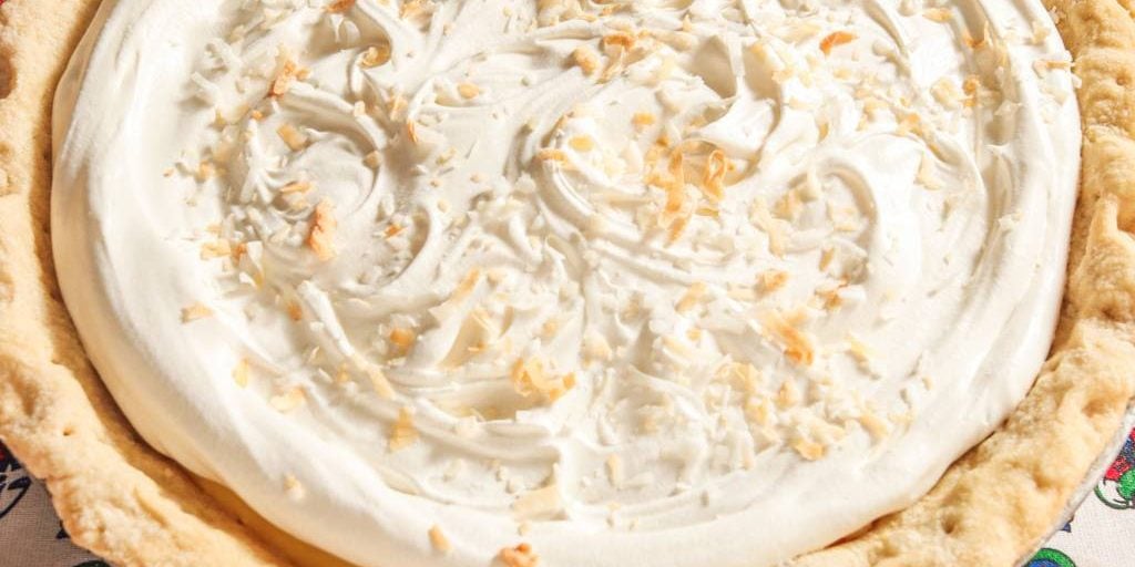 A close-up of a coconut cream pie with a golden, flaky crust, topped with swirls of whipped cream and sprinkled with toasted coconut flakes.