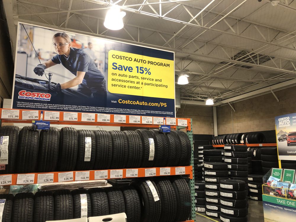 Display of stacked tires at Costco with a large yellow sign above promoting 15% savings on auto parts, service, and accessories through the Costco Auto Program. The ad also highlights popular Costco Products, featuring a woman working on a car.