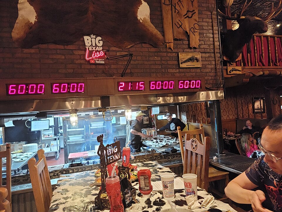A restaurant scene at The Big Texan with digital timers displaying 60:00, a central clock showing 21:15, and diners at a table with drinks and a large steak challenge display. Kitchen staff can be seen in the background.