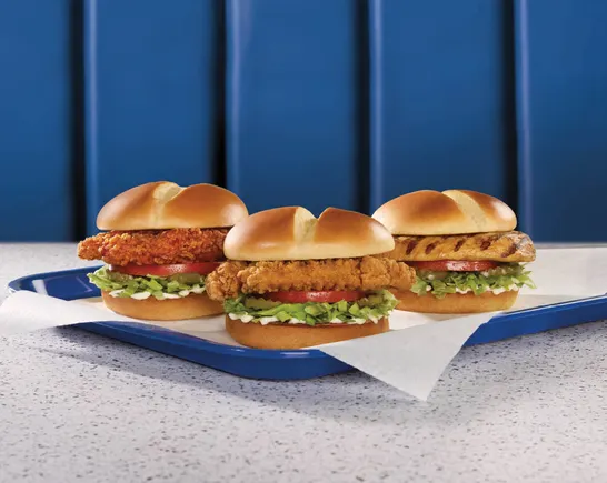 Three chicken sandwiches with lettuce and tomato are arranged on a blue tray lined with parchment paper, showcasing the classic Chicken Sandwich on a speckled countertop with a blue paneled wall in the background.