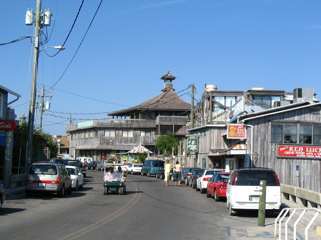 A street lined with parked cars and wooden buildings, including shops and restaurants. People walk and ride in a golf cart under a clear blue sky. A large round building with a central cupola stands at the end of the street.