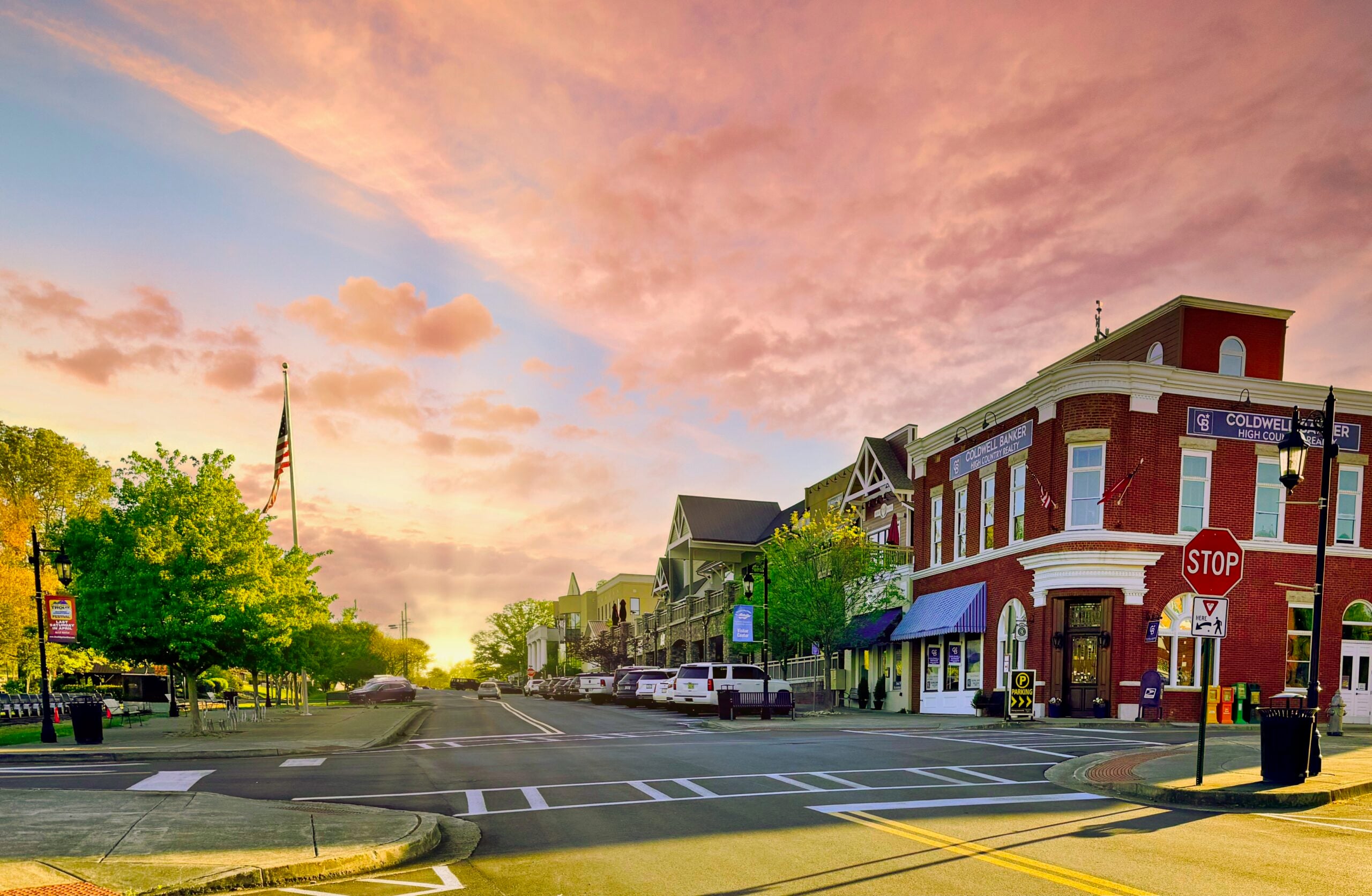 A small-town street at sunset with colorful clouds, a brick building housing a realty office, shops with awnings, a U.S. flag, trees, and a stop sign at the intersection. The street is quiet with no visible cars or people.