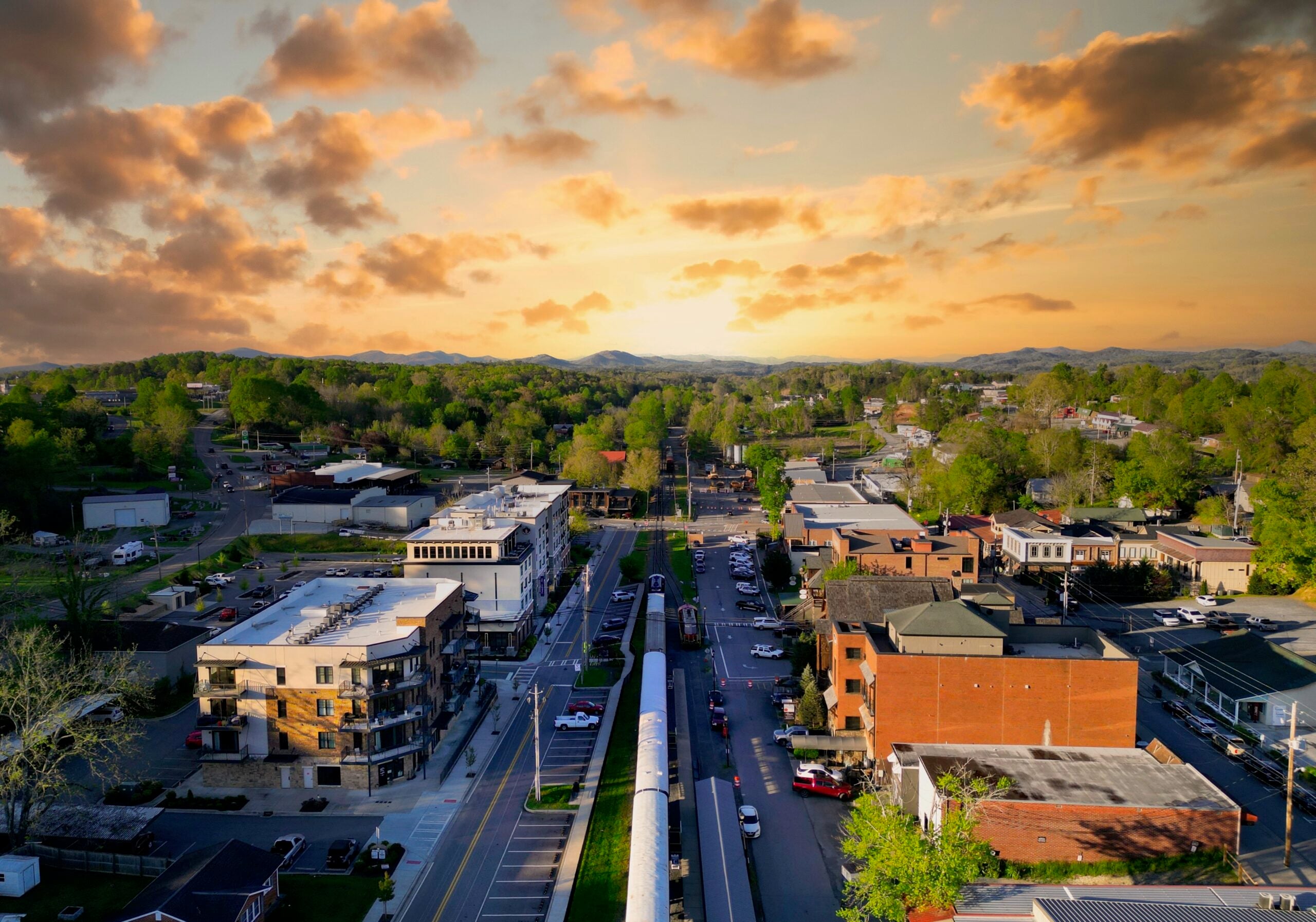 Aerial view of a small town at sunset with green trees, buildings, and cars lining the streets; a railway runs through the center toward a horizon of rolling hills under a colorful sky with scattered clouds.