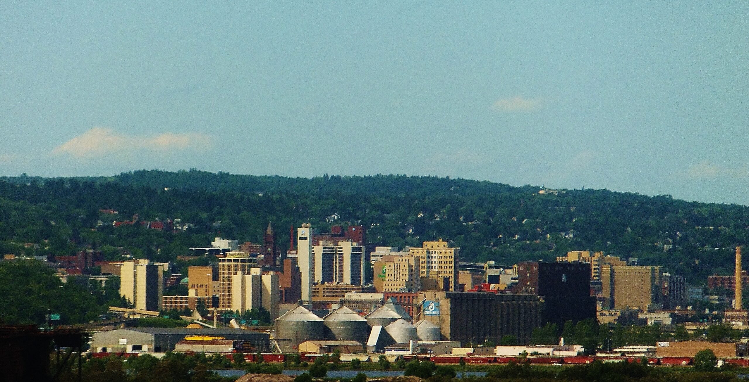 City skyline with various tall buildings and industrial silos in the foreground, set against a backdrop of green, tree-covered hills under a blue sky with a few clouds.