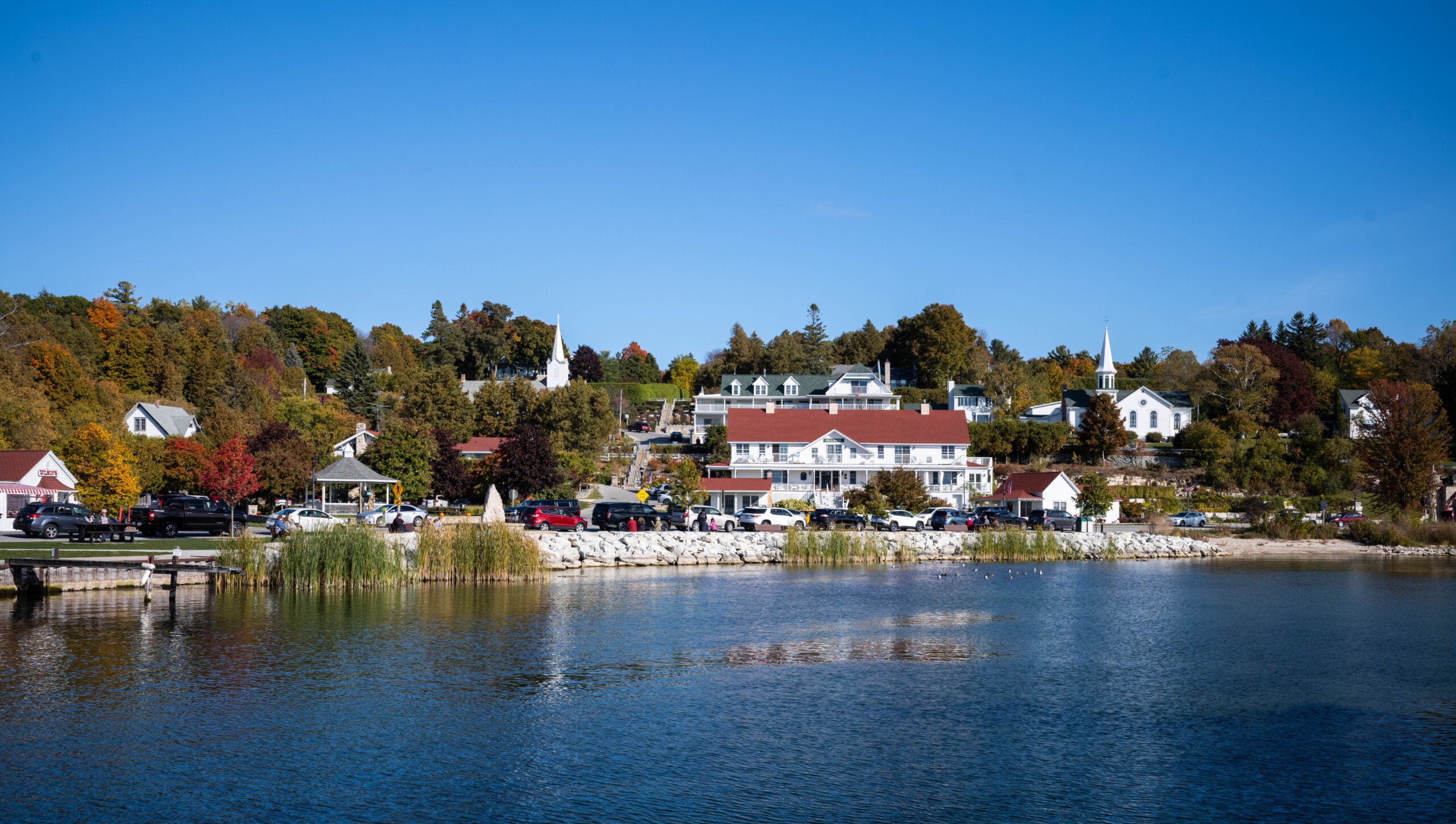 A scenic lakeside view of a small town with white and red-roofed buildings, trees in autumn colors, parked cars, and a clear blue sky reflected in the calm water.