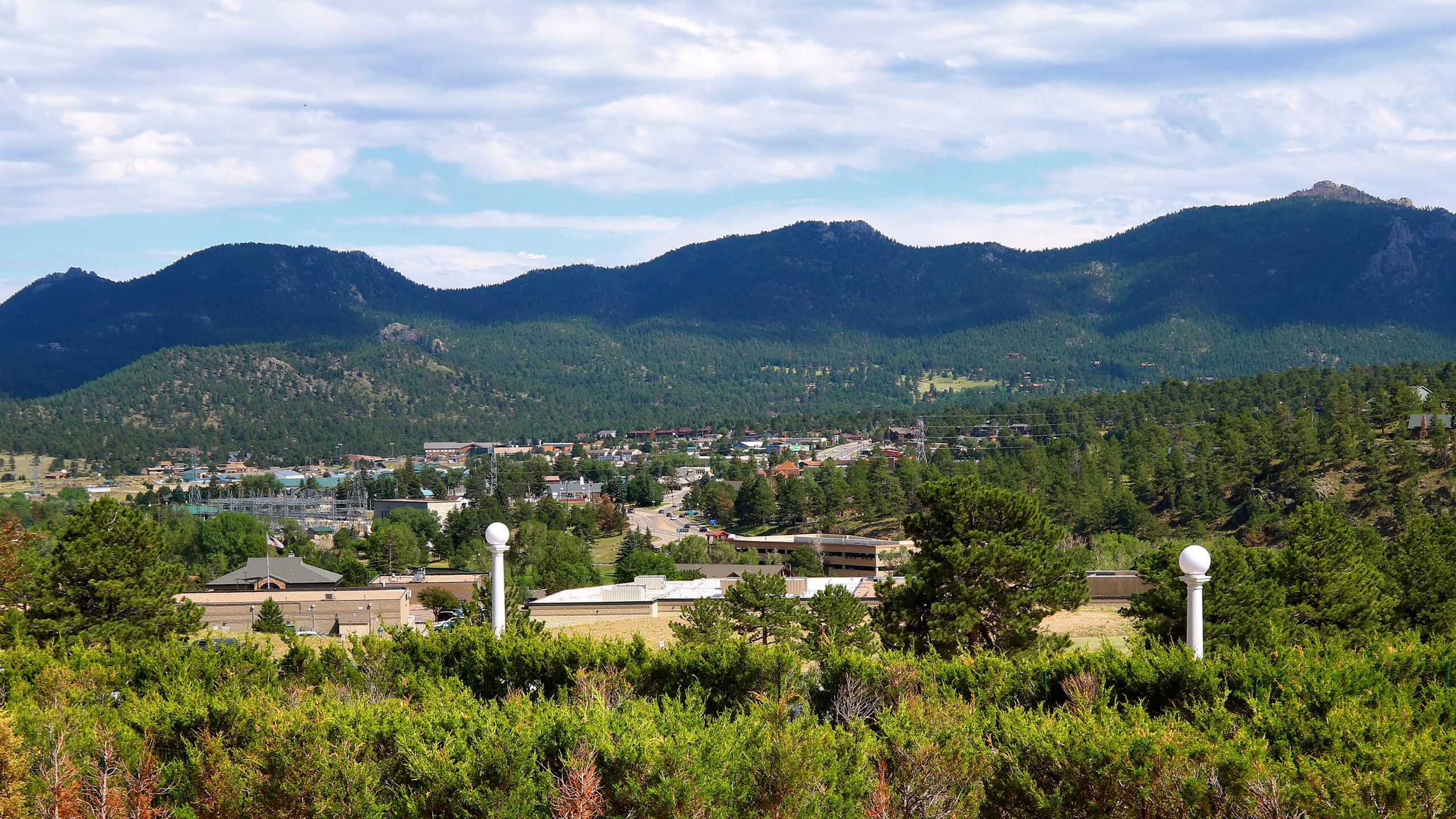 A scenic view of a small town nestled among green trees at the base of forested mountains under a partly cloudy sky. Two white lamp posts are visible in the foreground amid shrubs.