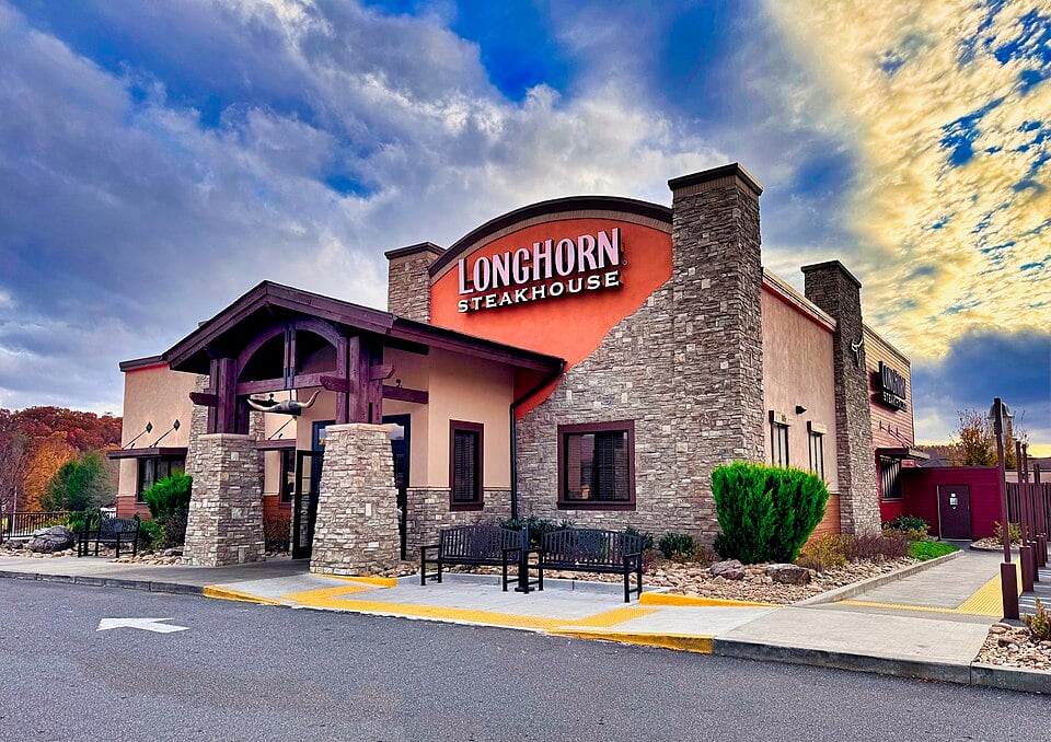 A LongHorn Steakhouse restaurant with a stone facade, large front entrance, and clear "LongHorn Steakhouse" signage under a dramatic, partly cloudy sky. Benches and shrubs line the walkway by the parking lot.