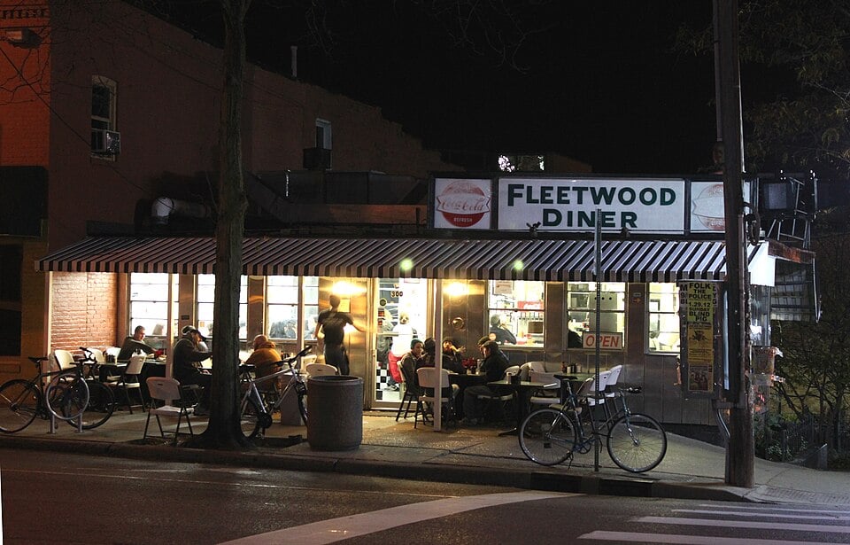A brightly lit diner called Fleetwood Diner is open at night, with people inside eating. Bicycles are parked outside along the sidewalk, and a striped awning covers the entrance. Trees and street signs are visible nearby.