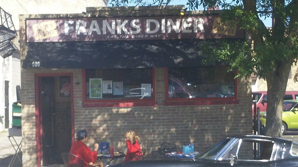 An outdoor view of Frank's Diner, a brick building with a black awning. Two people sit at a small table outside under a tree, and a classic black car is parked in front.