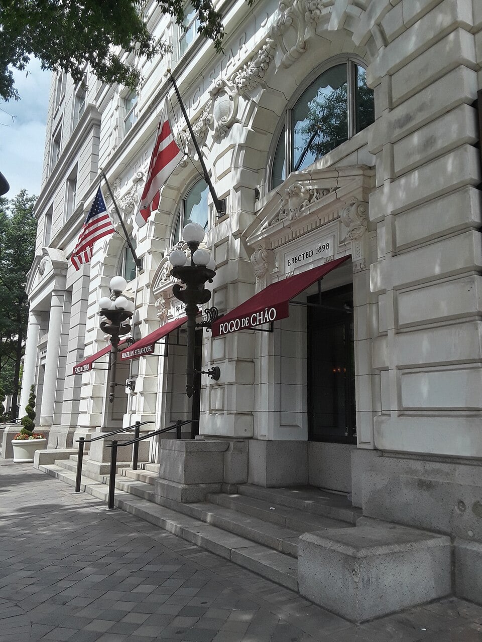 Restaurant entrance with red awning reading "Fogo de Chão," ornate white stone façade, two flags above (U.S. and another), decorative lamps, and arched windows, located on a city sidewalk.
