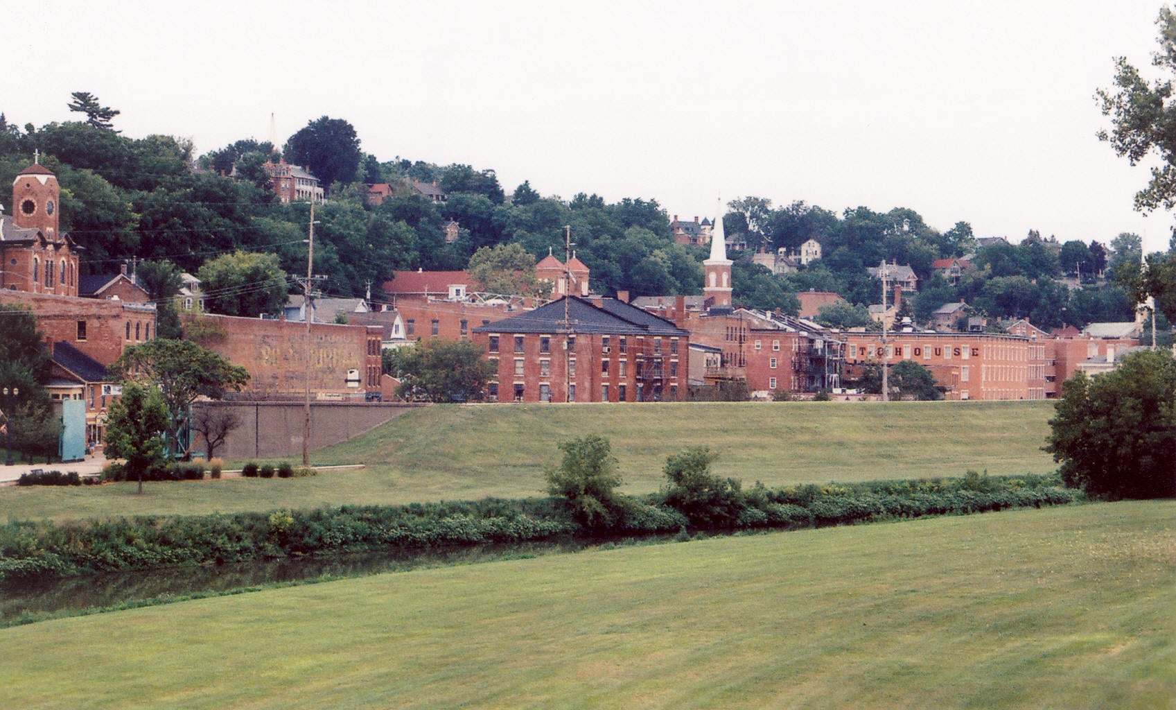 A scenic view of a small historic town with red brick buildings, steeples, and houses nestled on a green hillside, with an open grassy field in the foreground.