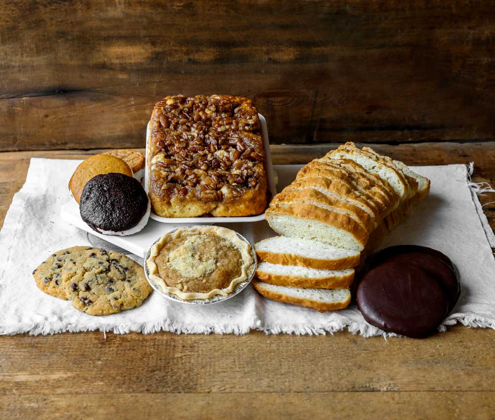 An assortment of baked goods on a white cloth, including sliced bread, cookies, a small pie, a sticky bun with nuts, a chocolate-covered treat, and ginger snaps, set against a rustic wooden background.
