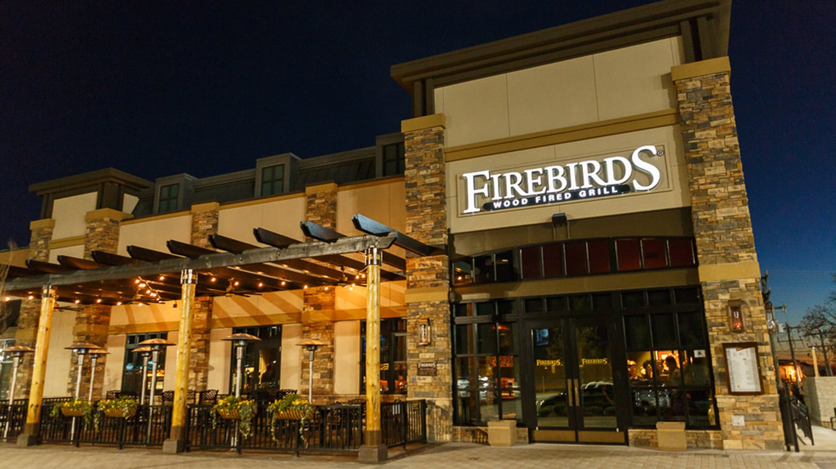 A large, well-lit restaurant with a sign reading “Firebirds Wood Fired Grill” above the entrance. The exterior features stone columns, outdoor seating, and large windows. It is nighttime.