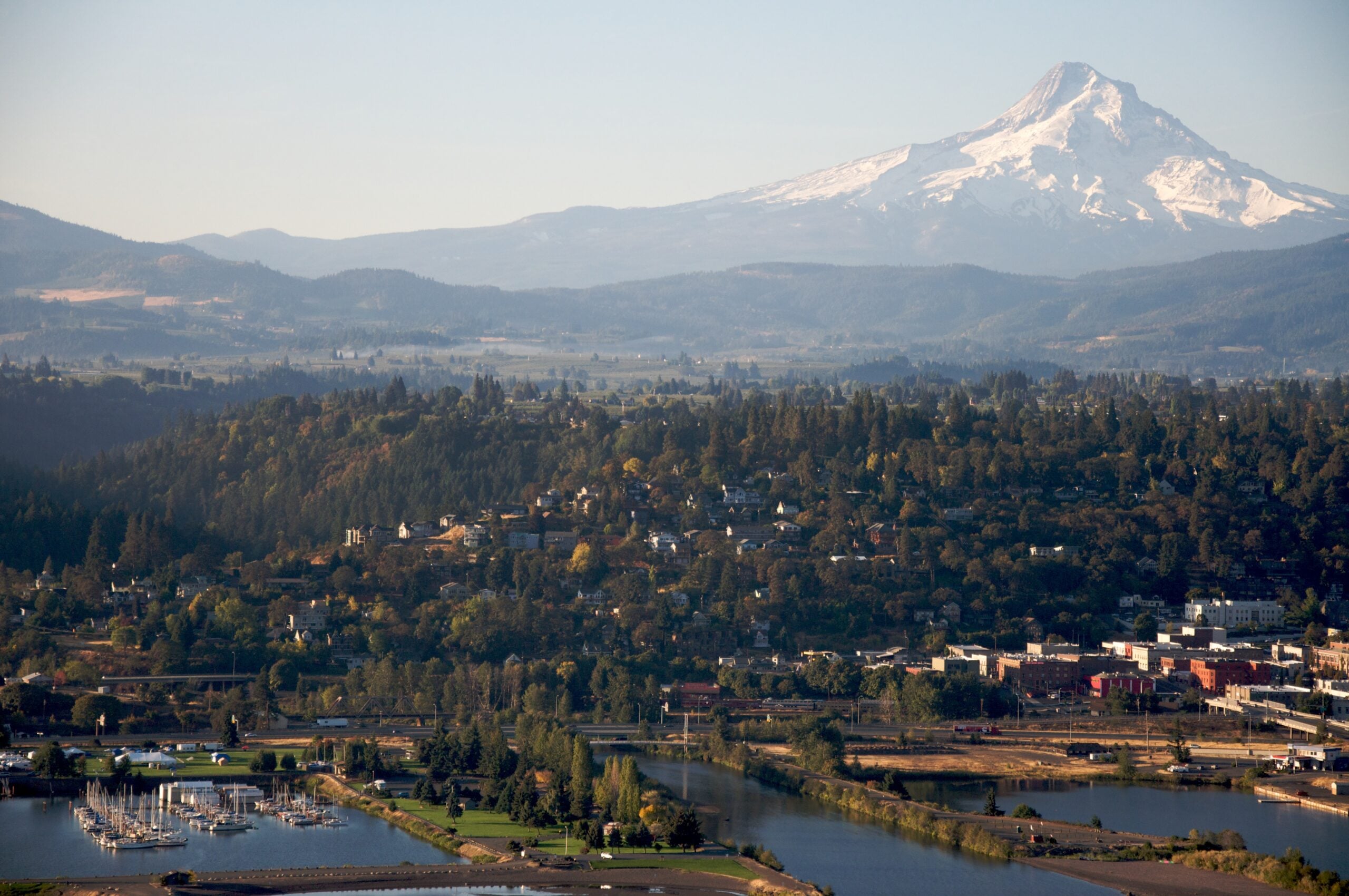 Aerial view of Hood River, Oregon, with a marina along the Columbia River, tree-covered hills, and the snow-capped peak of Mount Hood rising in the background under a hazy sky.