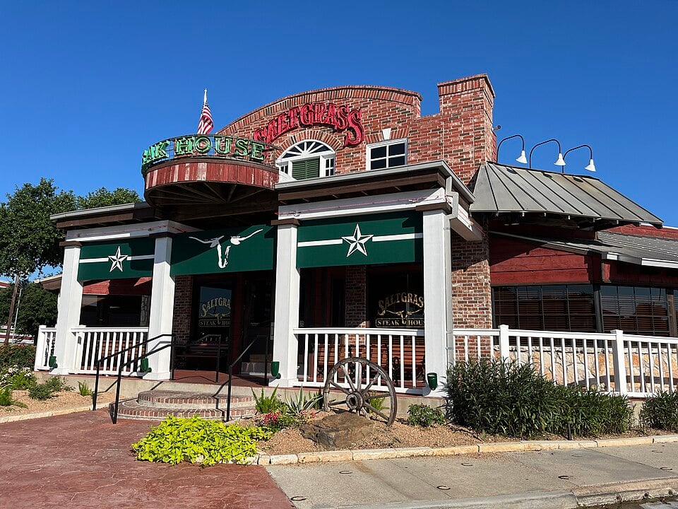 A brick and wood steakhouse with green awnings, white railings, and a western theme, including a wagon wheel in front; the sign reads "Saltgrass Steak House" under a clear blue sky.