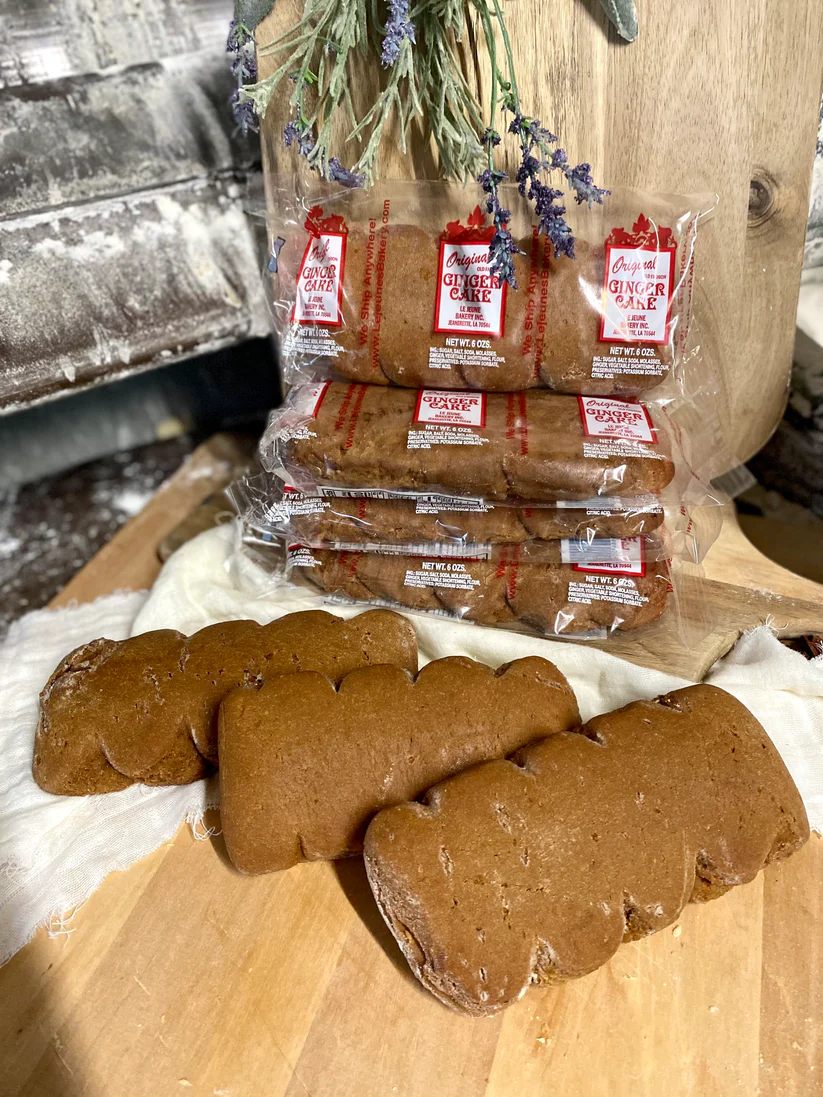 Three unwrapped rectangular ginger cakes and three stacks of plastic-wrapped ginger cakes are displayed on a wooden board, with dried lavender hanging above them and a rustic wooden backdrop.