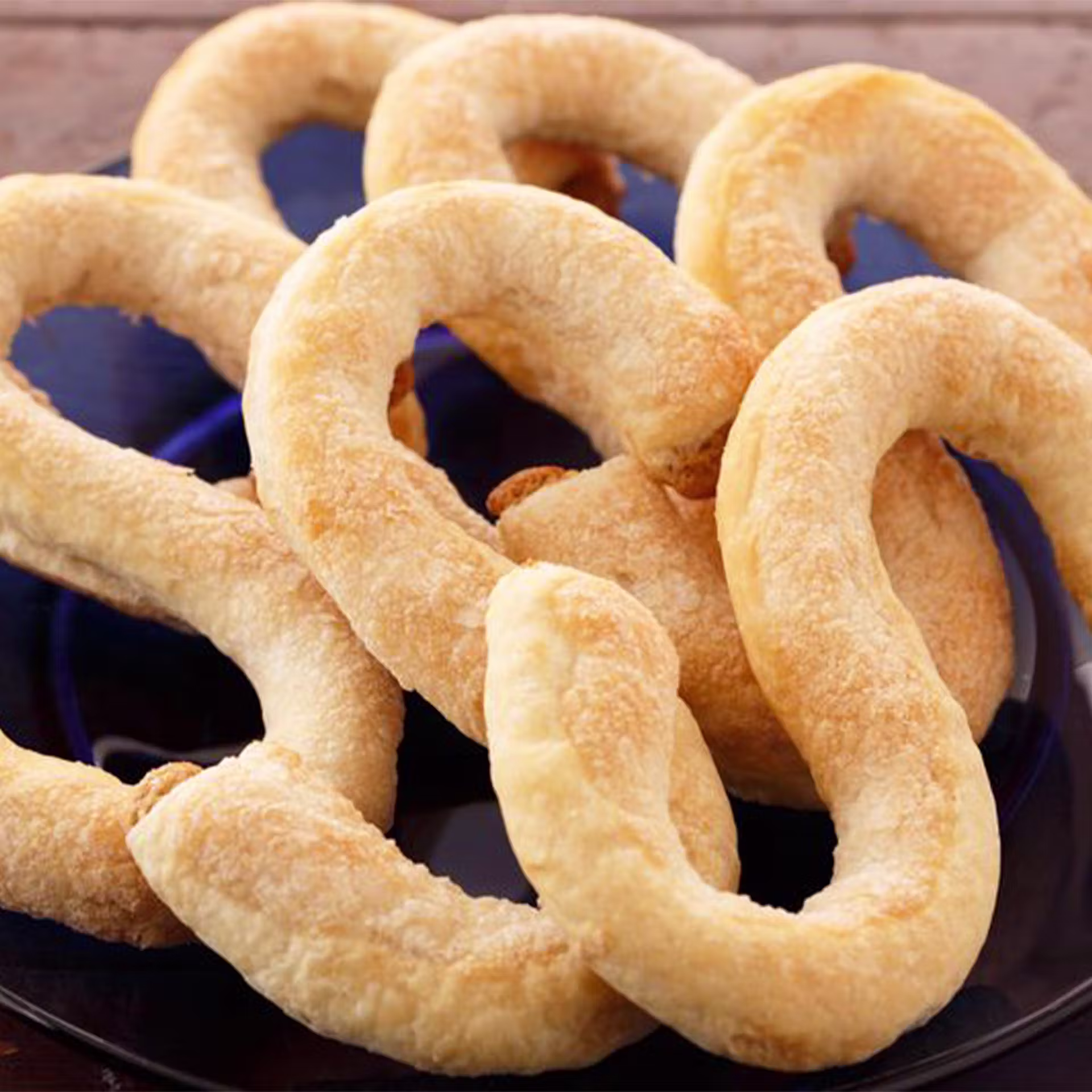 A black plate holds several S-shaped Cuban pastries called “rosquillas,” featuring a golden, flaky, and slightly rough texture, arranged so that some pieces overlap each other.