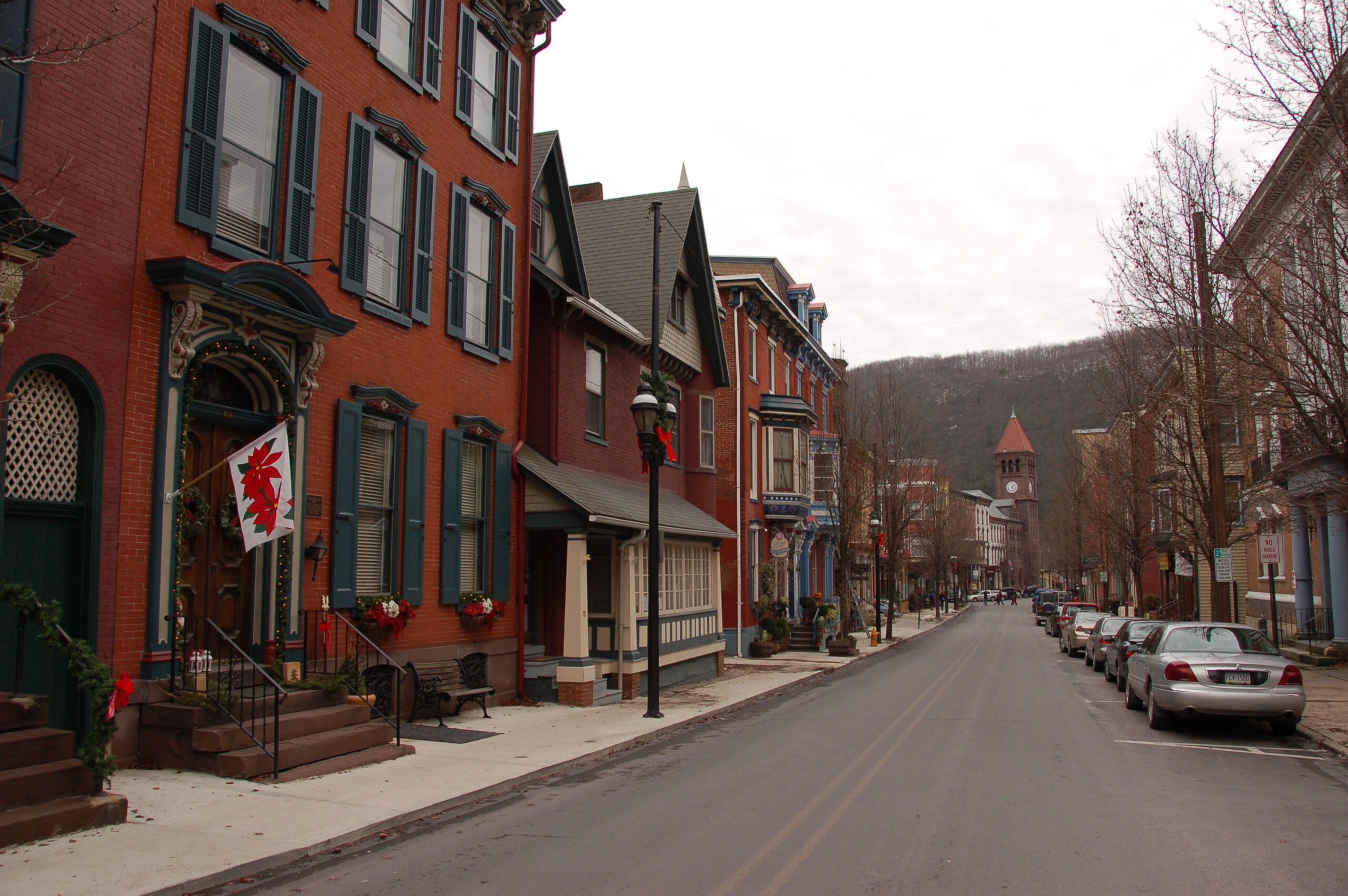 A quiet street lined with historic, colorful row houses and parked cars on a cloudy day. A red and white flag hangs from one house, and bare trees line the sidewalk. A church steeple is visible in the distance.
