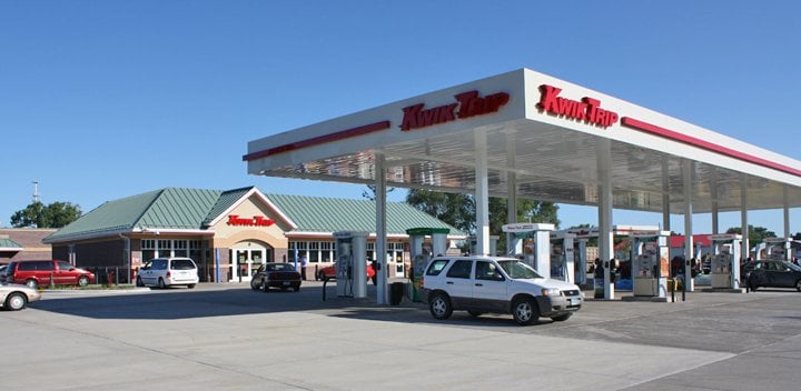A Kwik Trip gas station and convenience store with multiple fuel pumps, a few cars parked by the pumps, and a green-roofed building in the background under a clear sky.