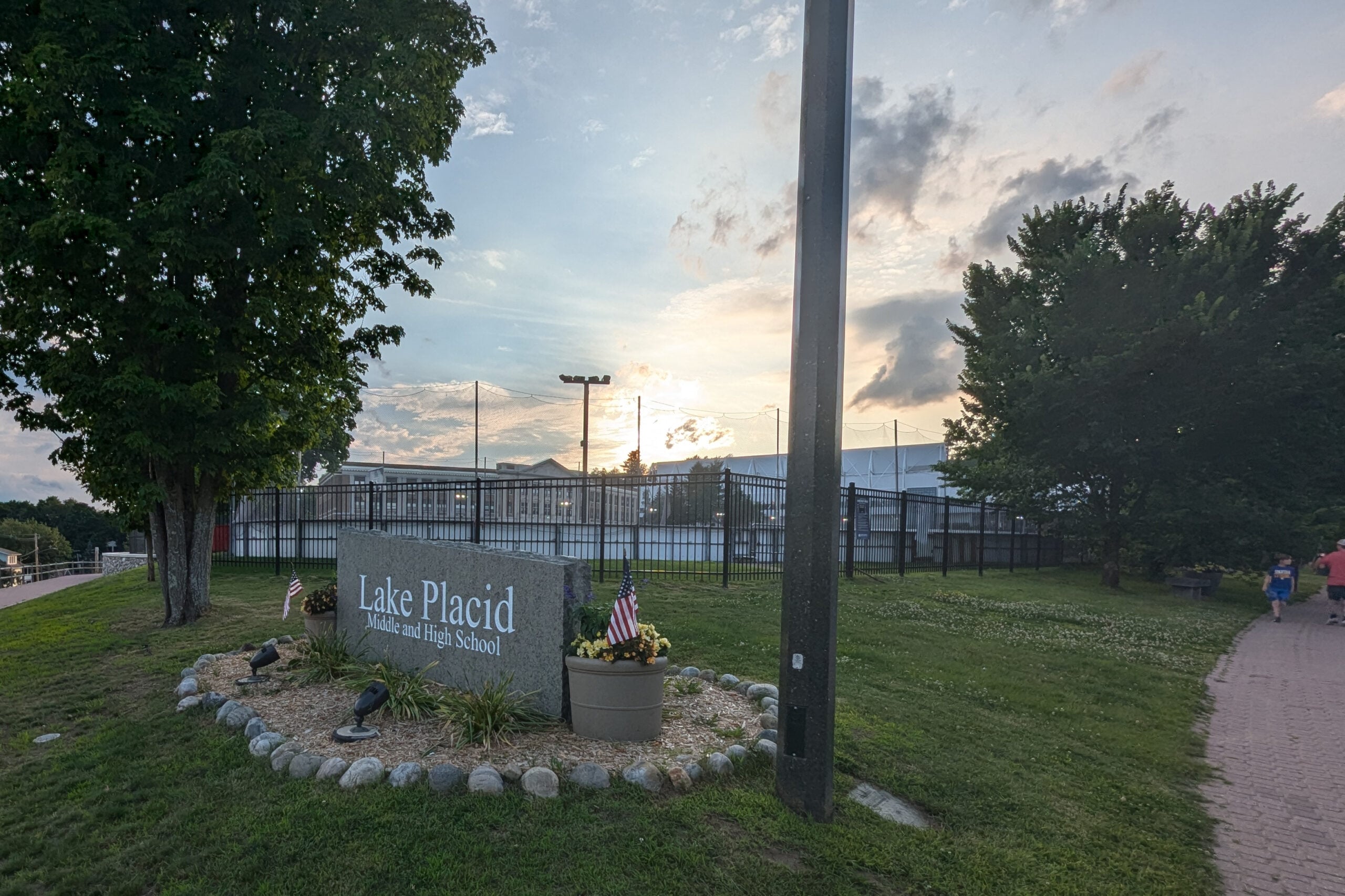 A stone sign reading "Lake Placid Middle and High School" sits near a sidewalk, surrounded by grass, trees, and small American flags, with school buildings and a cloudy sky in the background at sunset.