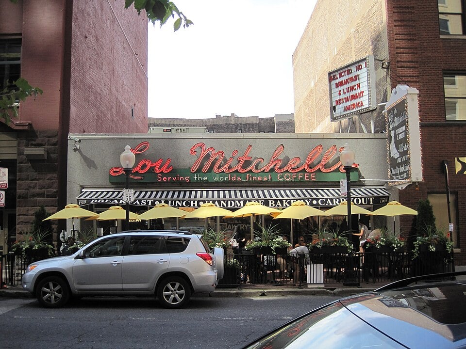 Lou Mitchell’s restaurant exterior with a retro neon sign, yellow umbrellas shading outdoor tables, and people dining on a city sidewalk. A red SUV is parked in front, with nearby buildings visible in the background.