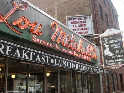 A neon sign for Lou Mitchell’s restaurant reads “Serving the world’s finest COFFEE.” Additional signs advertise breakfast, lunch, and handmade bakery goods on a brick building.