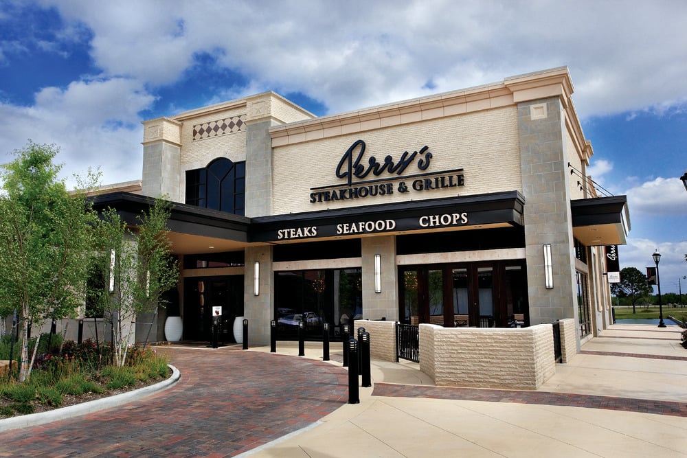 The exterior of a modern steakhouse restaurant named "Perry's Steakhouse & Grille," with large windows, stone walls, and signs advertising steaks, seafood, and chops under a partly cloudy sky.