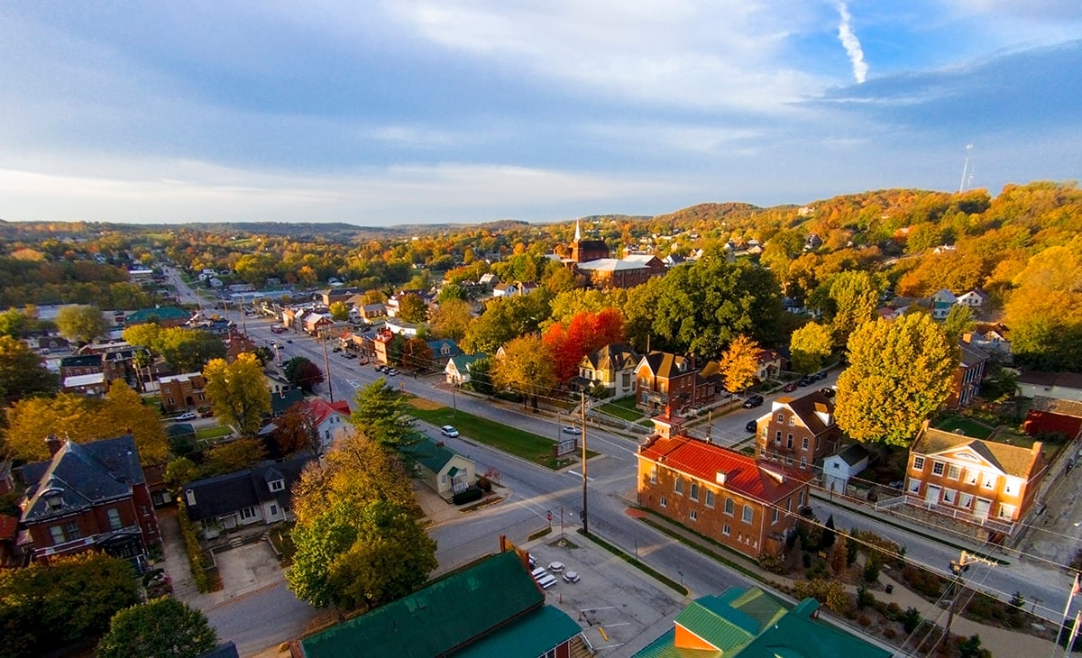 Aerial view of a small town with tree-lined streets, colorful autumn foliage, and a mix of historic and modern buildings under a partly cloudy sky.