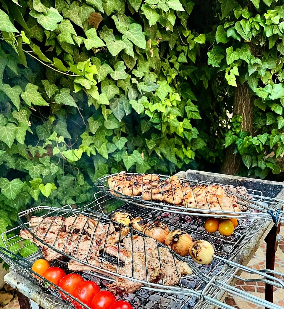 Skewers of grilled meat, cherry tomatoes, and onions cooking on a barbecue grill, with a leafy green ivy background.