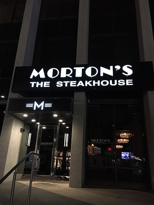 Nighttime view of the entrance to Morton’s The Steakhouse, featuring a black awning with illuminated white lettering and a glowing "M" logo by the door.