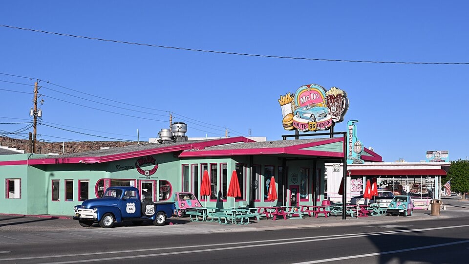 A retro-style diner with mint green and pink decor, outdoor seating, red umbrellas, and vintage cars parked outside. A large sign reads “Mr. D’z Route 66 Diner” against a clear blue sky.