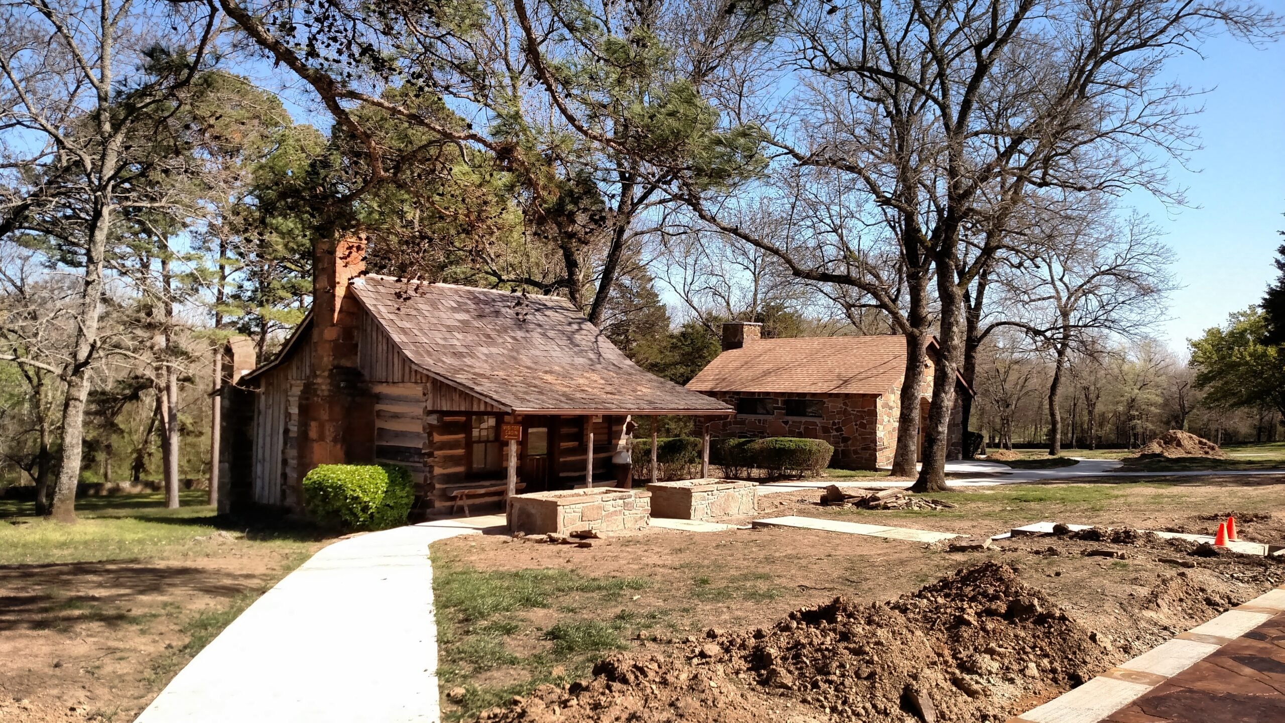 A rustic log cabin and a stone house sit among leafless trees on a sunny day. Winding concrete paths lead to the buildings, and there is some dug-up ground in the foreground. The area is surrounded by grass and trees.