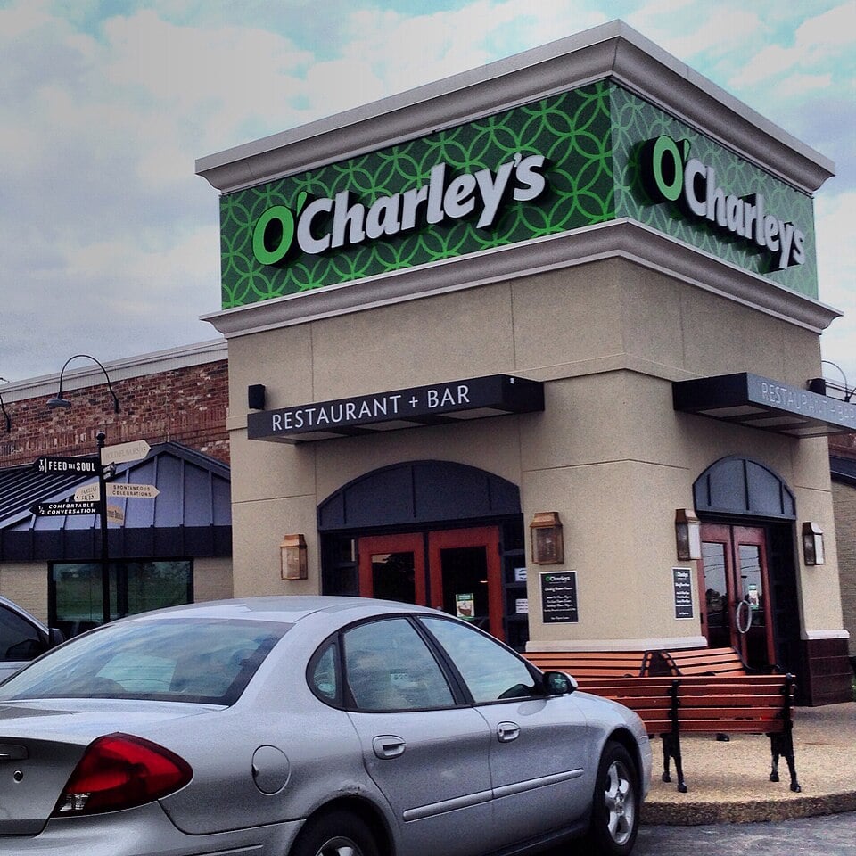 A silver car is parked in front of an O'Charley's restaurant and bar; the building has a green patterned sign and beige walls. The entrance has benches and wall lanterns.
