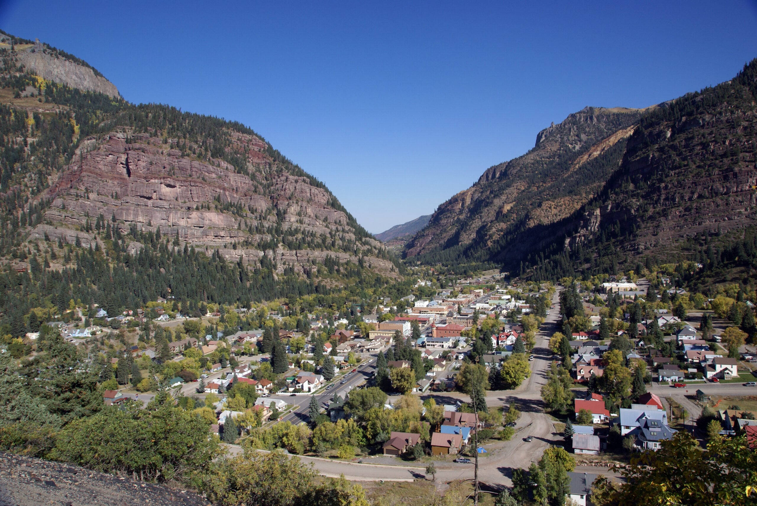 A small town is nestled in a green valley between two rocky, forested mountains under a clear blue sky. The town has a mix of houses, roads, and trees, surrounded by dramatic, rugged cliffs.
