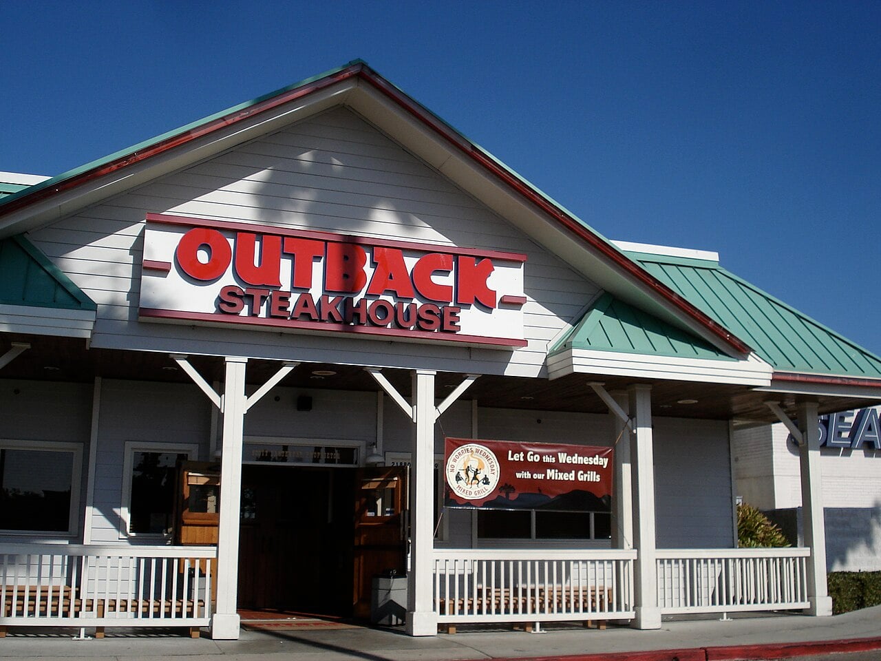 Outback Steakhouse exterior with a green metal roof, white walls, and a large red and white sign above the entrance. A banner on the porch advertises Wednesday specials. The sky is clear and blue.