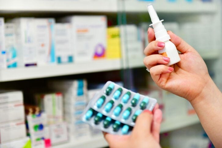 A person holds a nasal spray in one hand and a blister pack of capsules in the other, standing in front of shelves filled with various medications at a pharmacy.
