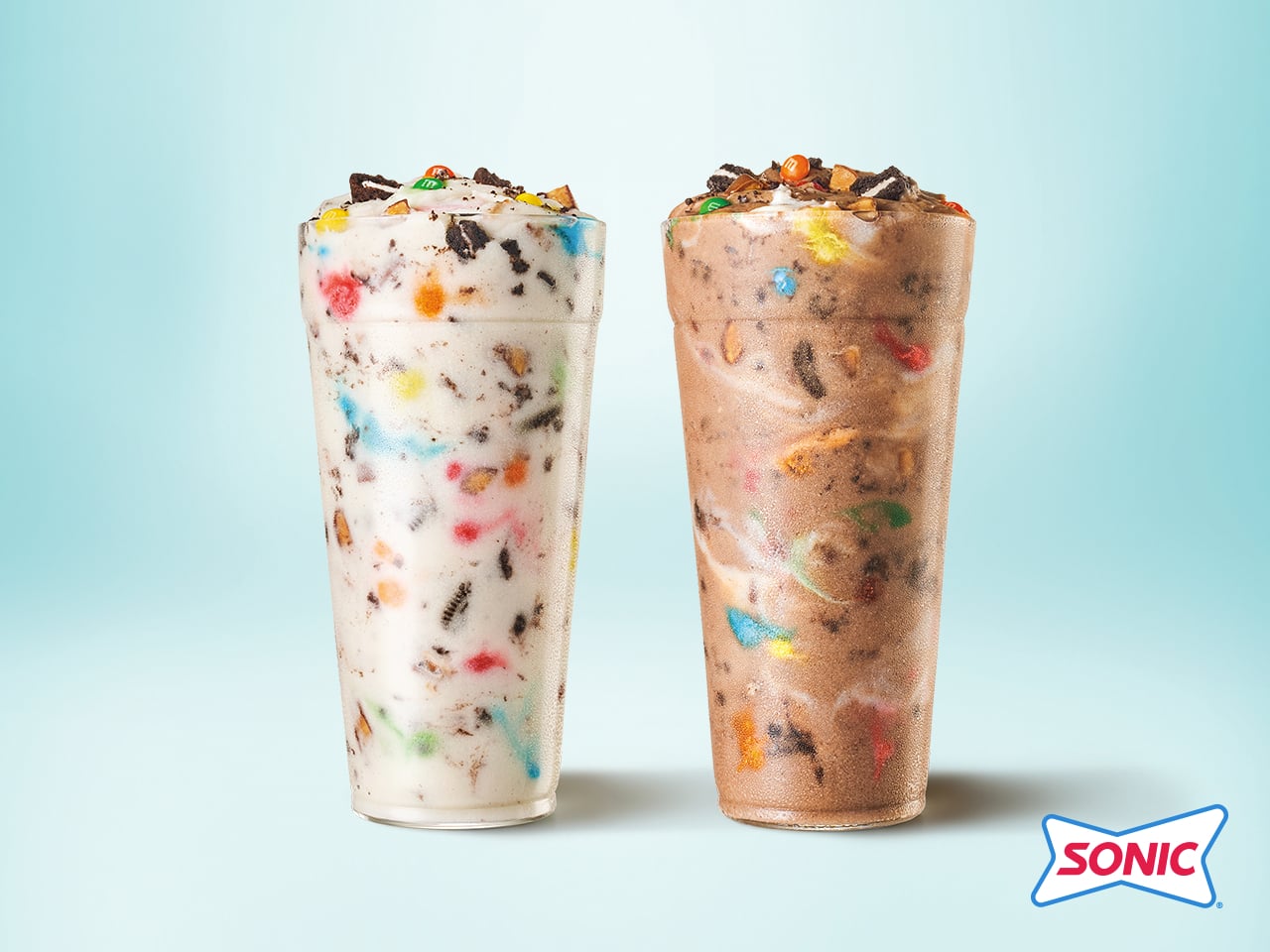 Two tall Sonic cups filled with creamy blended fast-food desserts, one vanilla and one chocolate, both mixed with colorful candy pieces and cookie bits, against a light blue background with the Sonic logo in the corner.