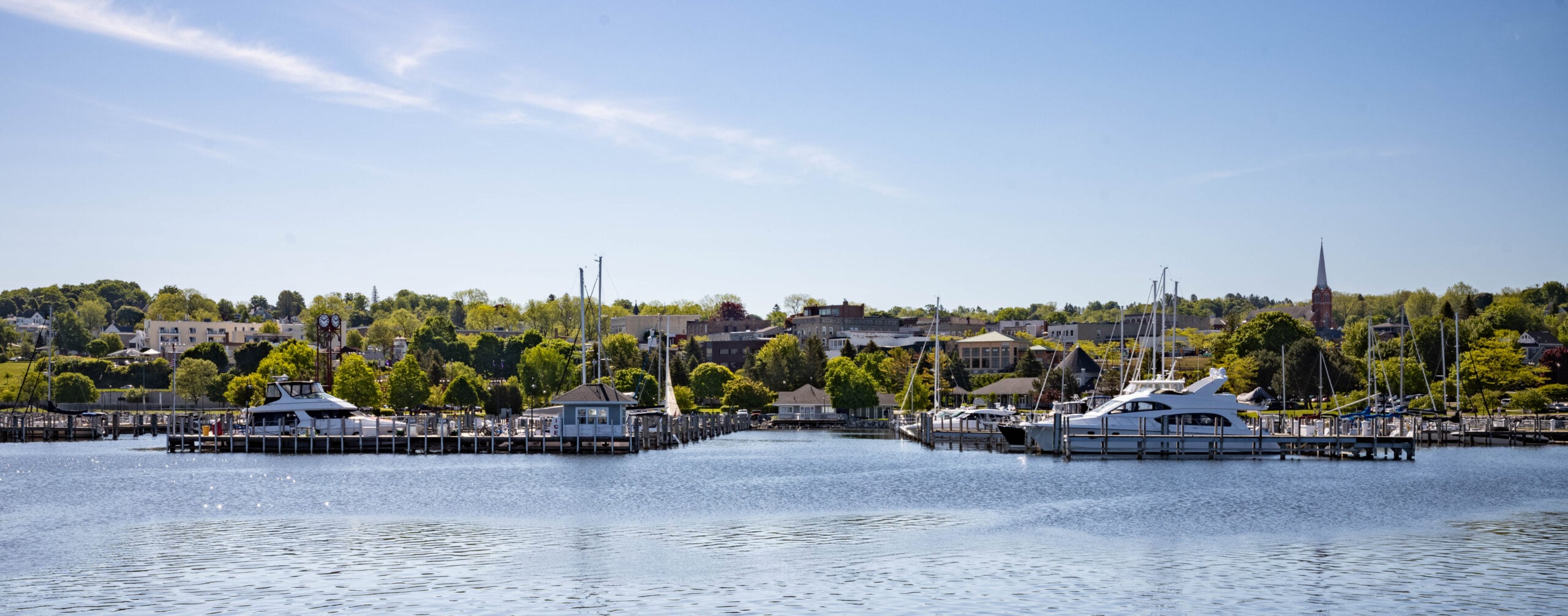 A calm marina with several boats docked at wooden piers, set against a background of green trees, buildings, and a church steeple under a clear blue sky.