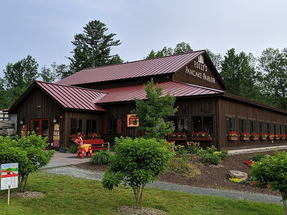 A rustic wooden building with a red metal roof, labeled “Polly’s Pancake Parlor,” is surrounded by greenery and flower boxes, with trees and a cloudy sky in the background.