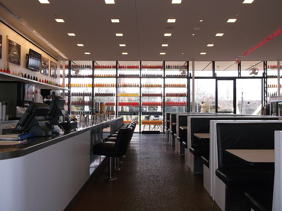 A modern diner with black-and-white booths and bar stools, a counter with registers, and large windows lined with colorful bottles. The restaurant is empty and well-lit with ceiling lights.