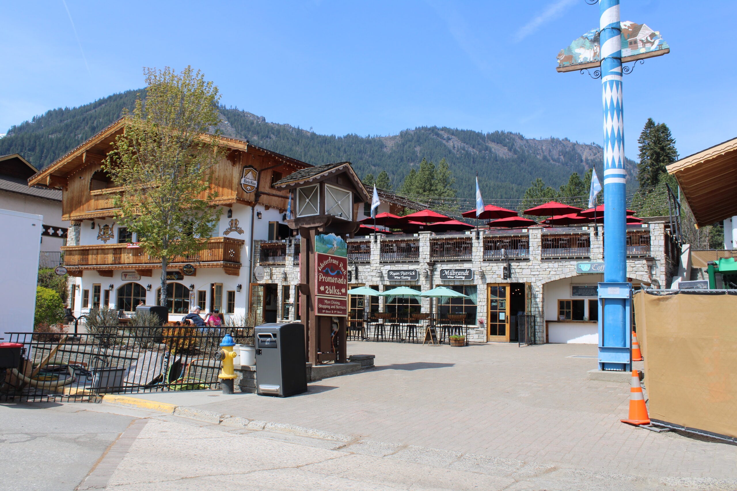 A Bavarian-style building with wooden balconies and a restaurant patio with red umbrellas, set against a backdrop of forested mountains under a blue sky. A tree, streetlamp, and sidewalk are in the foreground.