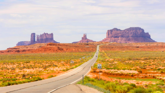 A long, straight road winds through a desert landscape toward large red rock formations under a partly cloudy sky—a perfect scene that inspires thoughts of taking vacations more often. A “Speed Limit 65” sign stands beside the road in the foreground.