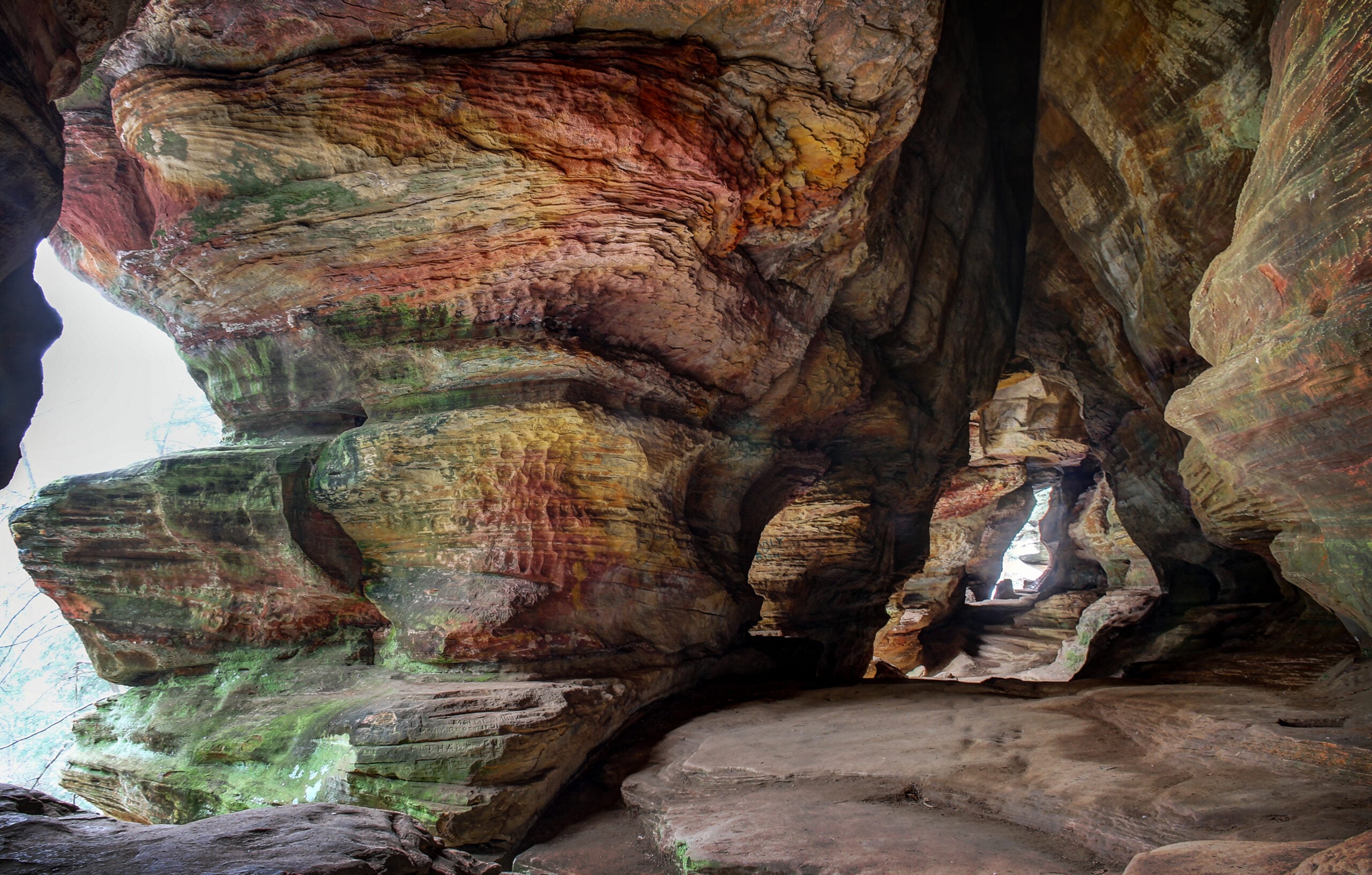Large, colorful rock formations inside a cave, with layered walls of red, yellow, and green hues. Natural light enters through an opening at the end of a winding, rocky passageway.