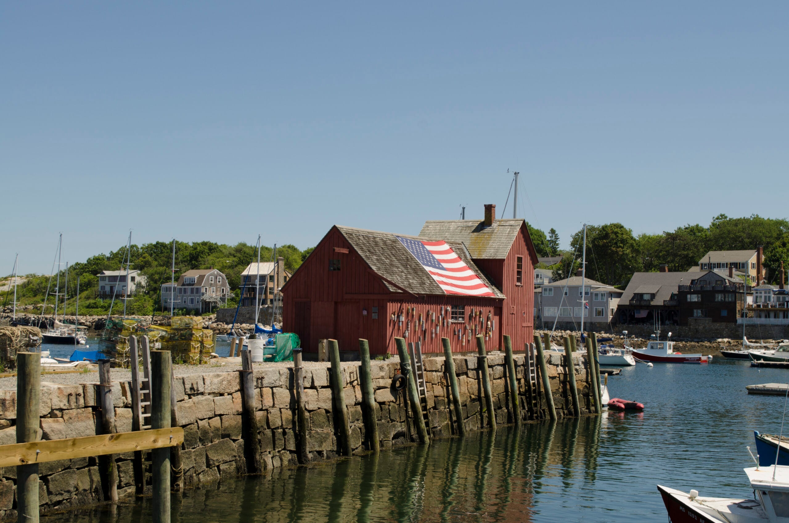 A red wooden building with an American flag draped over its roof sits on a stone pier by the water, surrounded by boats and waterfront houses under a clear blue sky.