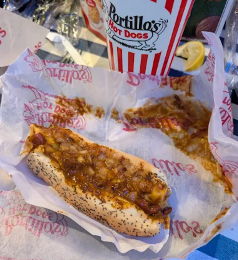 A chili cheese dog in a poppy seed bun sits on Portillo’s branded paper, with some spilled chili next to it—a classic fast-food hot dog. A large striped Portillo’s Hot Dogs cup and a lemon slice are in the background.