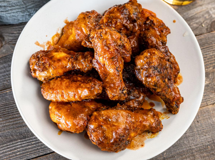 A white plate filled with several crispy, saucy chicken drumsticks and wings, coated in a reddish-brown glaze of flavorful wing sauces, sitting on a rustic wooden table.