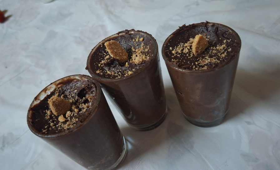 Three glasses filled with thick chocolate mousse, each topped with crumbled biscuit pieces, are arranged on a light-colored tablecloth—a favorite treat enjoyed by middle class moms during family gatherings.