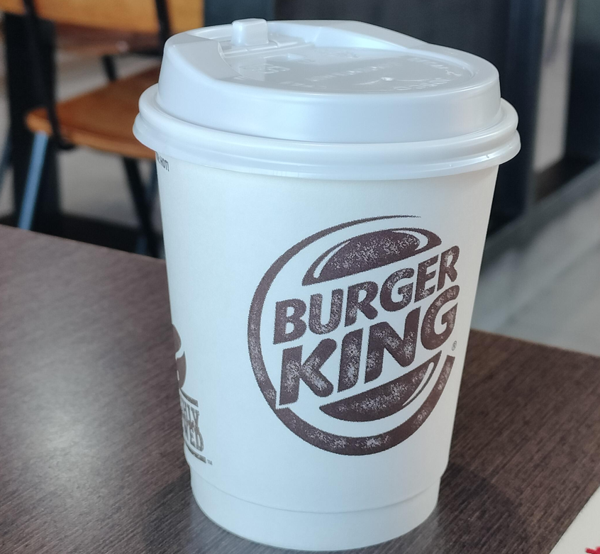 A white Burger King takeaway coffee cup with a plastic lid sits on a dark brown table in a restaurant setting, showcasing the appeal of fast food coffees amid chairs and a blurred background.
