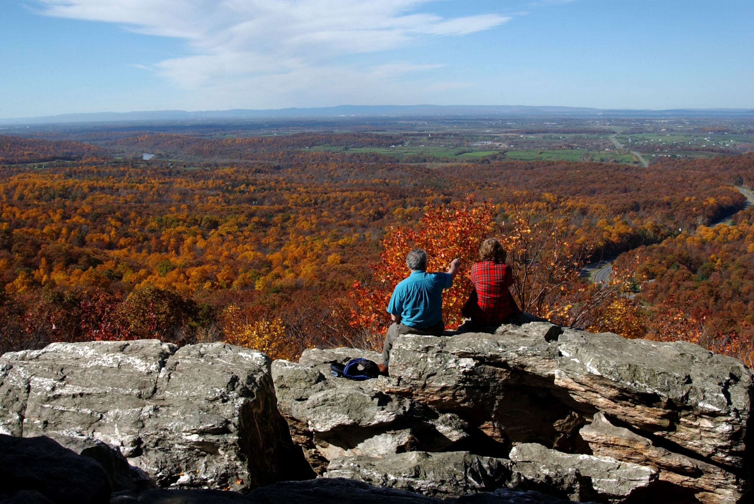 Two people sit on a rocky ledge overlooking a vast forest with autumn foliage. One person points toward the colorful trees and distant river under a blue sky with wispy clouds.